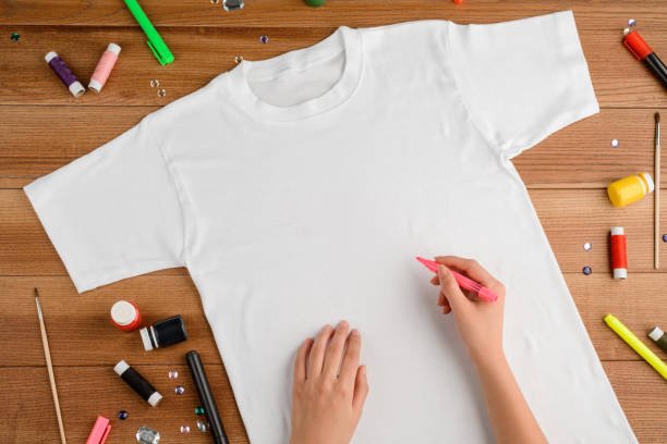 Girl draws on a t-shirt with a fabric marker, copy space. Wooden table with paint, rhinestones and threads. Makeover concept.